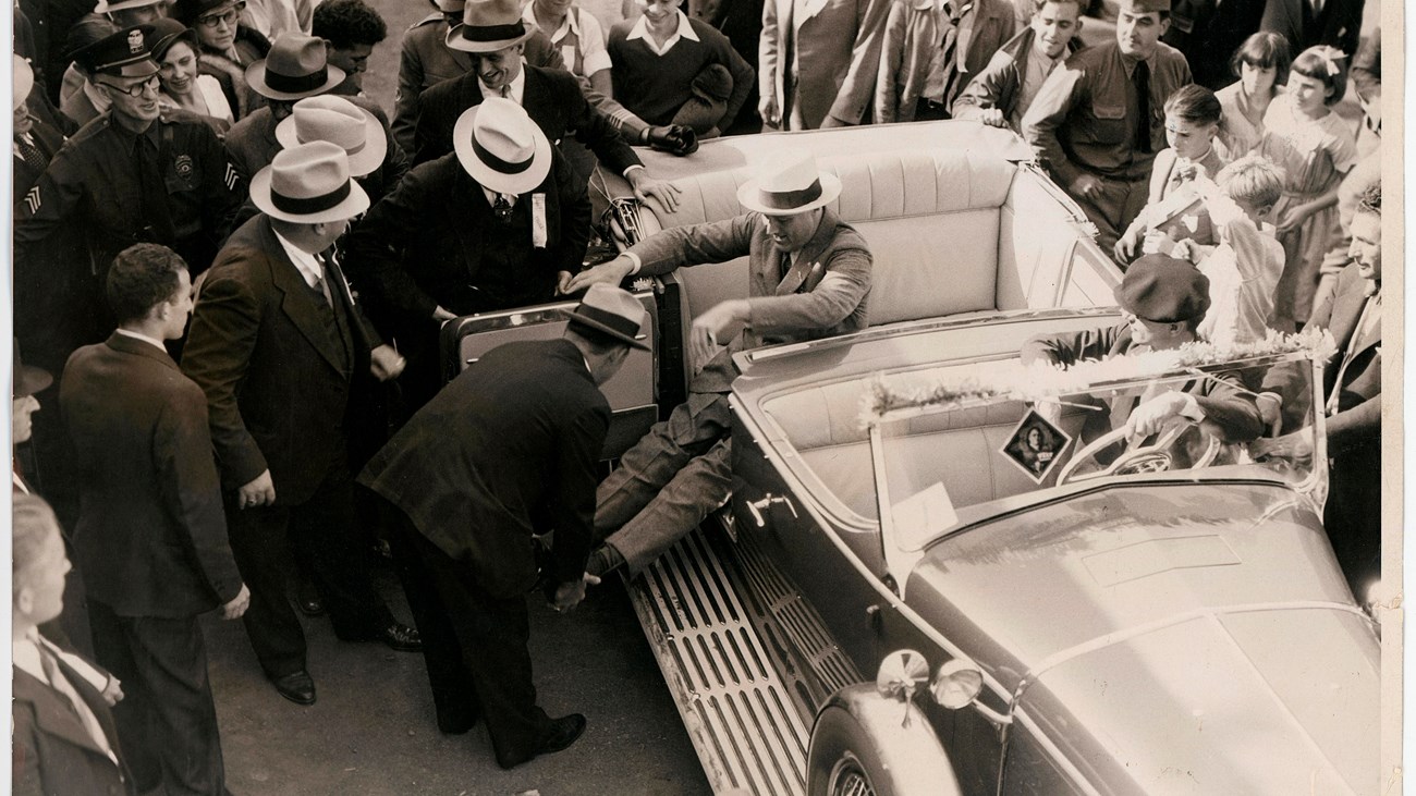 An eager crowd watches as Secret Service agents straighten and lock FDR’s leg braces in preparation 