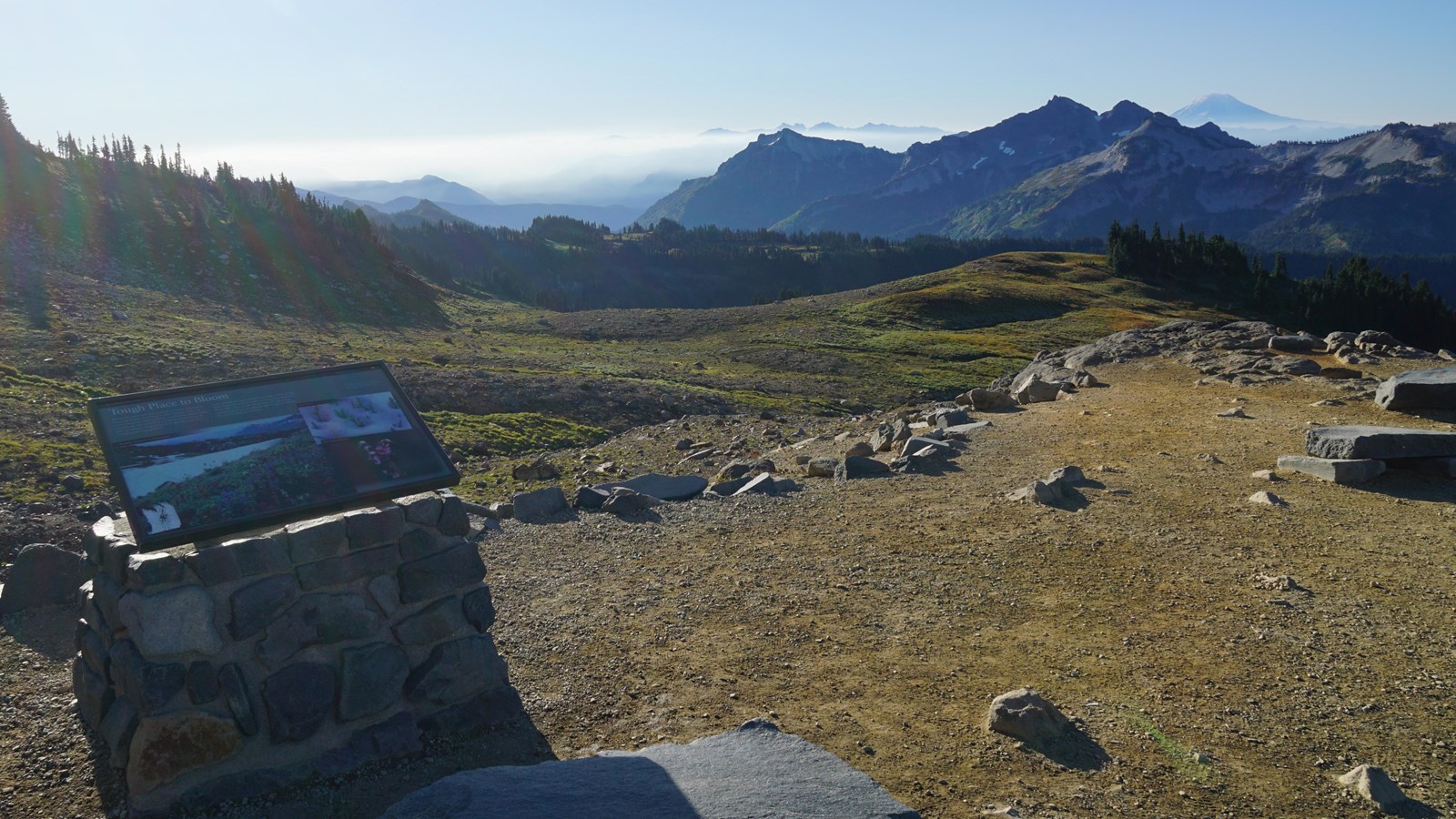 A wayside panel attached to a rock stand overlooking a meadow and distant mountain peaks.