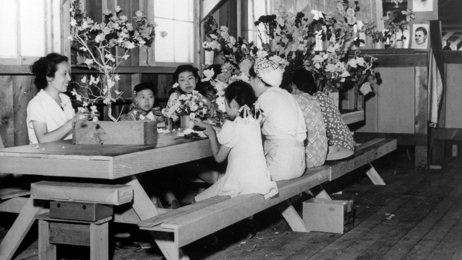 Women and girls sit on a wooden picnic table inside a building creating flower arrangements