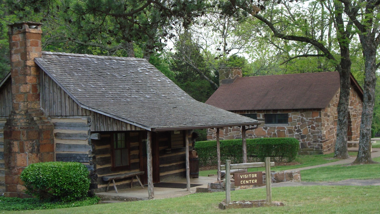 A historic, small log cabin, with an awning, sits in front of a distant small stone cabin in forest.