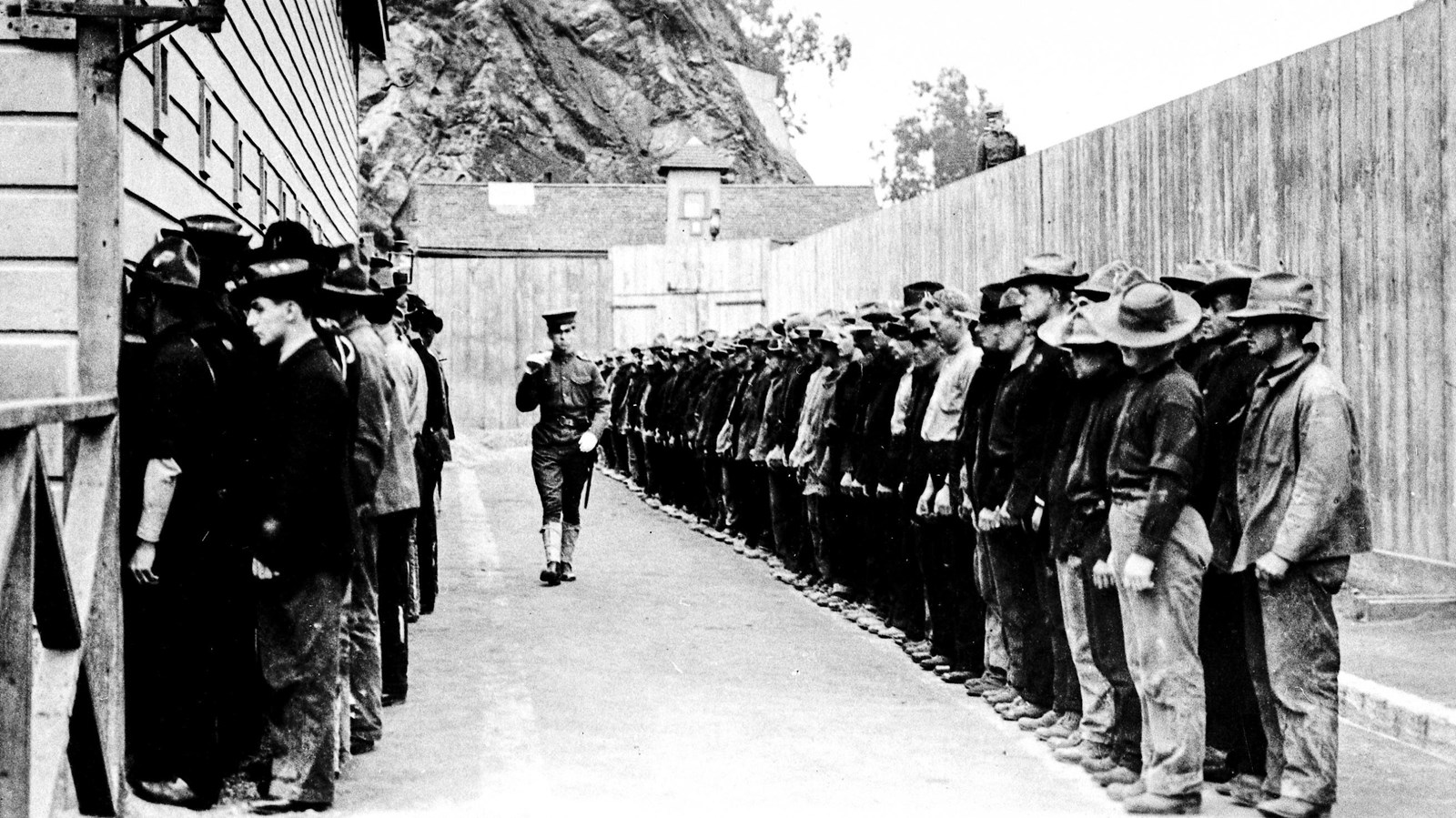 A guard walks past prisoners on either side of him, lined up in two long rows.