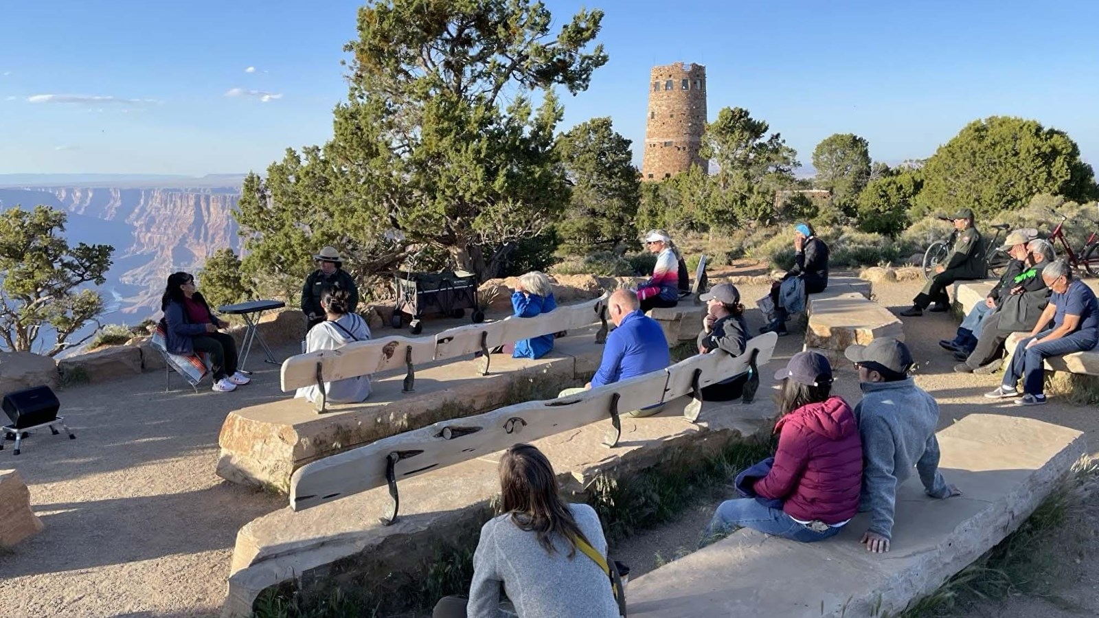 15 people are sitting on stone slab benches in an outdoor amphitheater on the edge of the canyon.