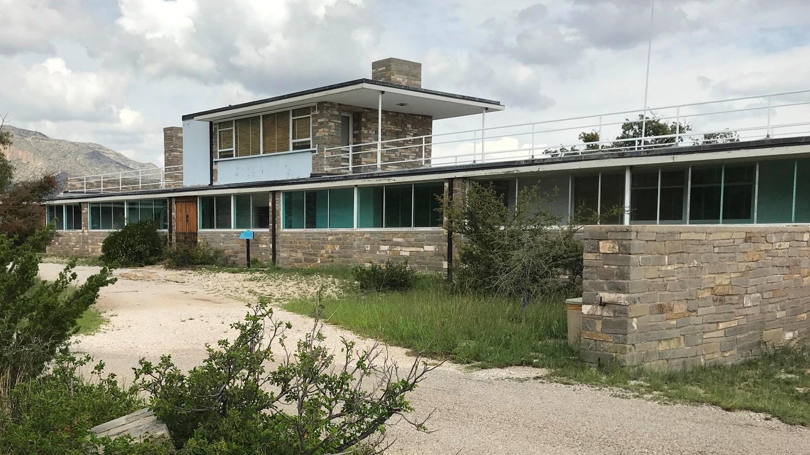 A rectangular house made of yellow stone rises in front of mountains.