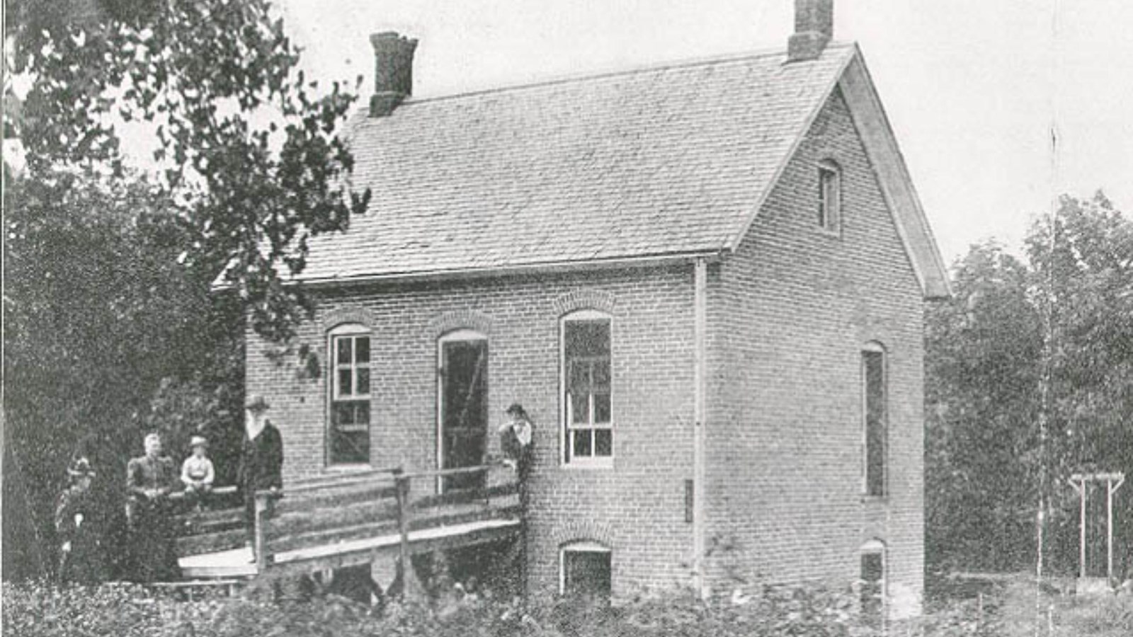 People stand on a small bridge leading into a brick home. Black and white image.