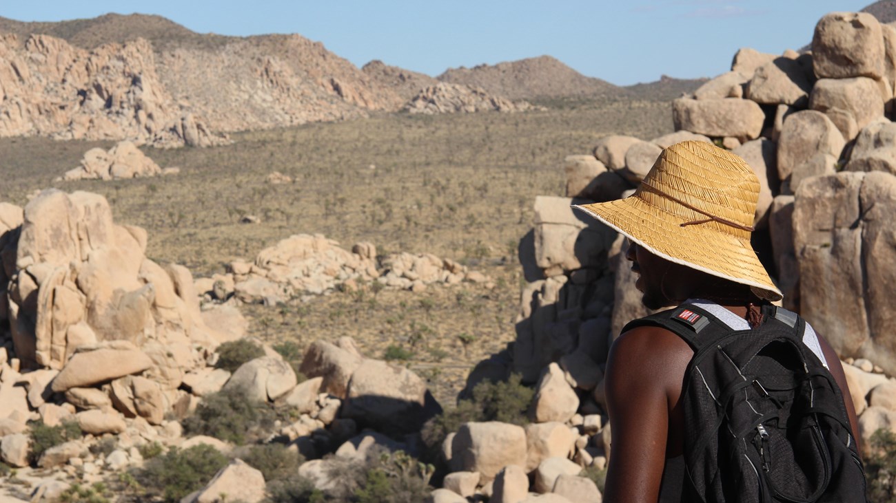 A hiker looking through a rocky landscape to a valley with mountains in the distance.