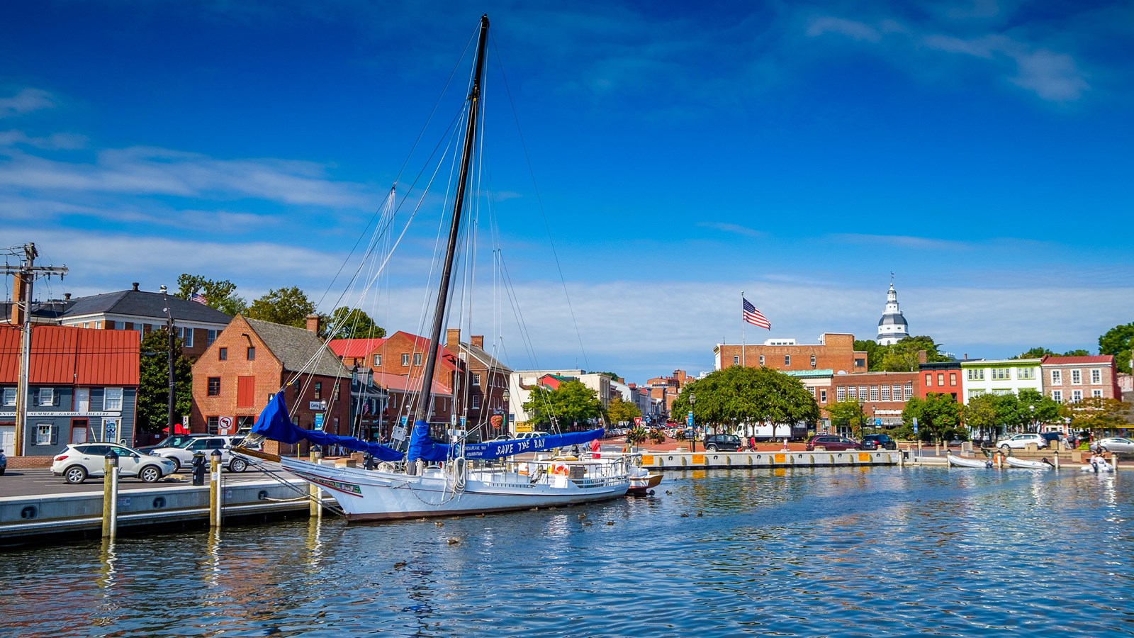 White sailboat floats in front of brick buildings and green trees.