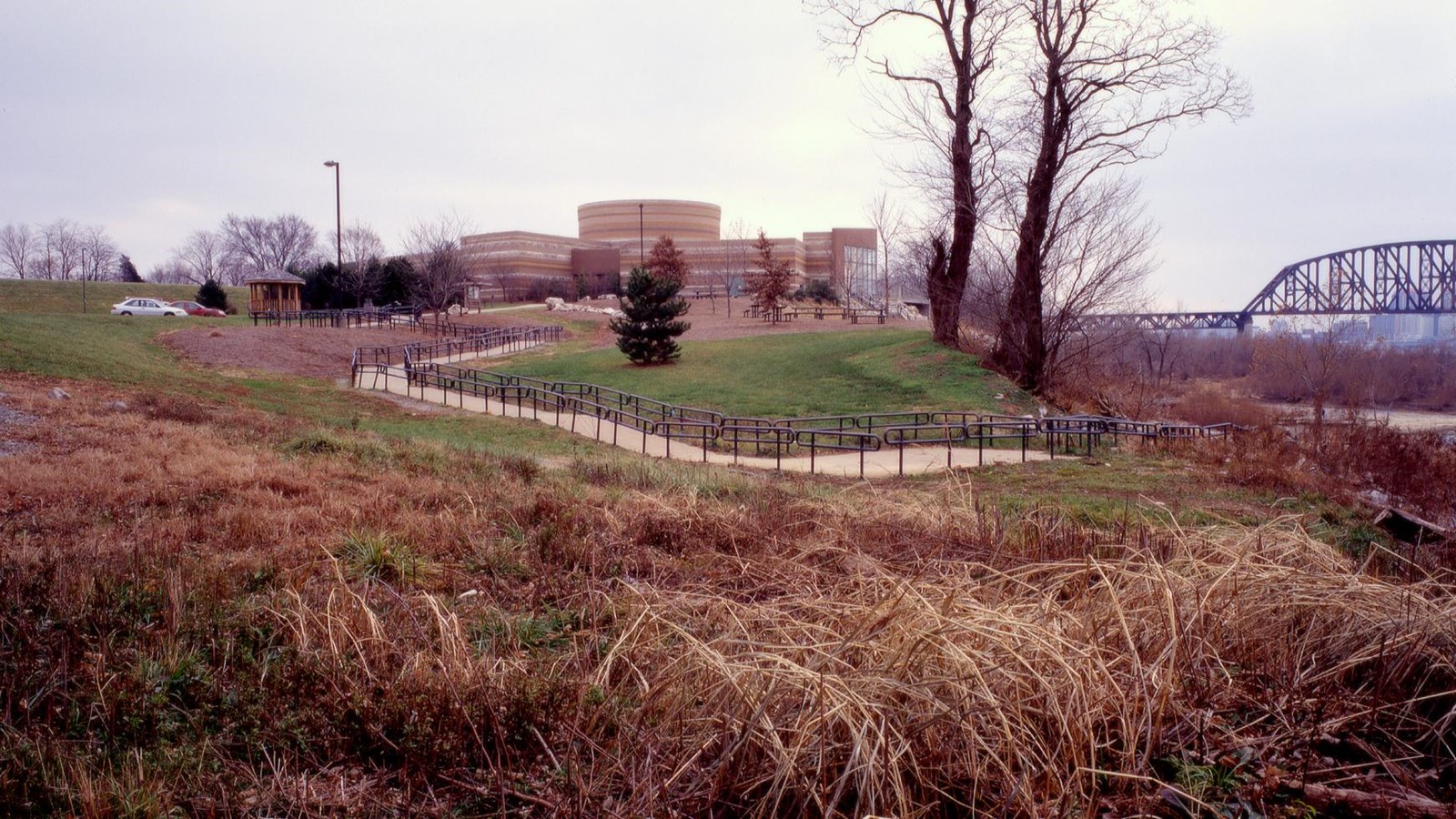 A multi-level brick building sits amid a green grassland. A trail winds down to the river