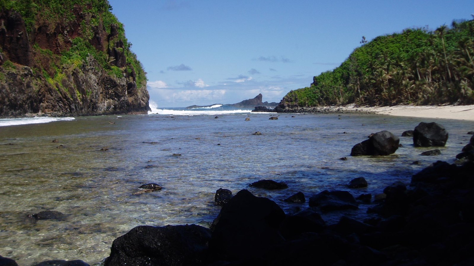 A view of a rocky shore and wide body of water with green trees on both sides against a blue sky.