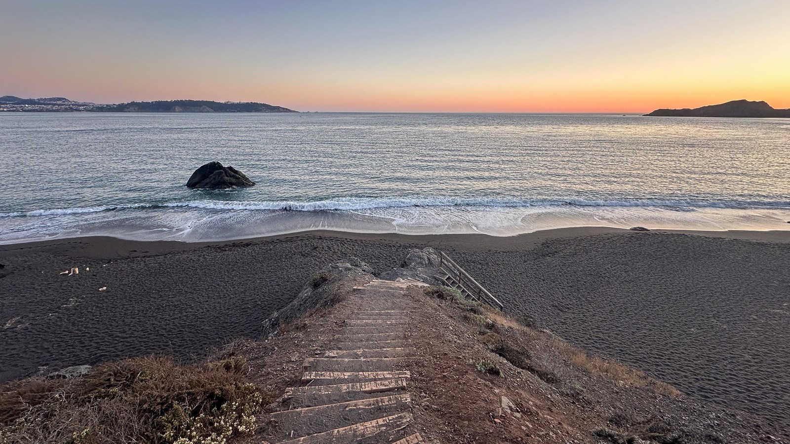 Wooden stairs descend toward a beach with black sands at sunset.