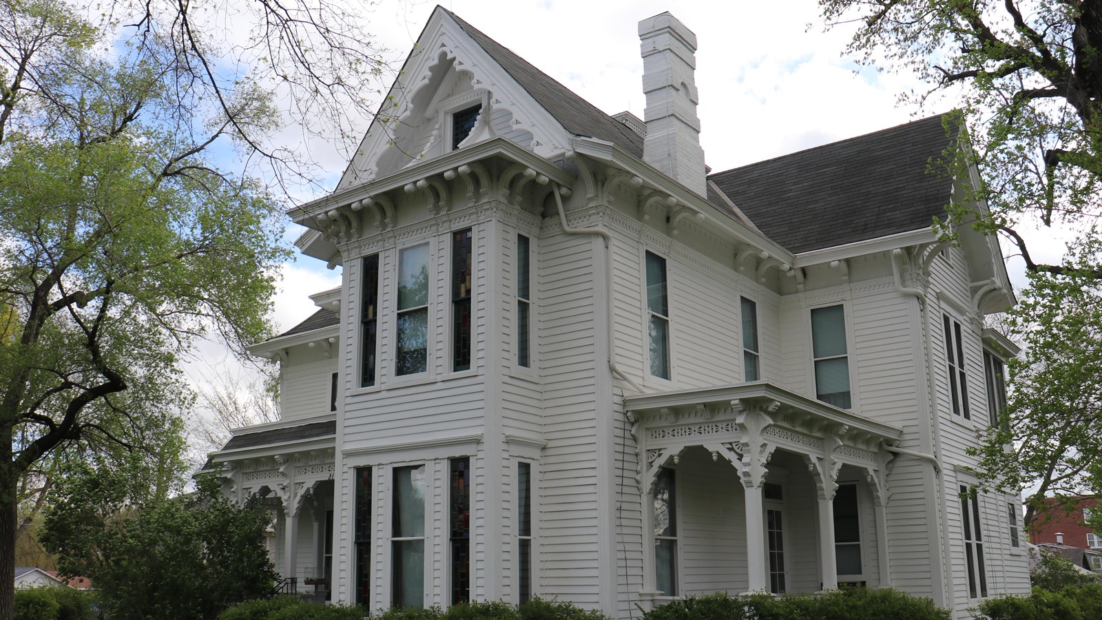 A white two and a half story Victorian home with gingerbread woodwork on the two porches with bushes