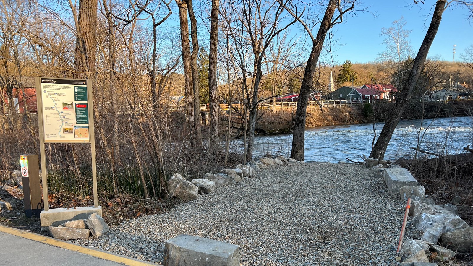 A gravel path lined with large rocks and trees leads to the river. To the left is an information pan