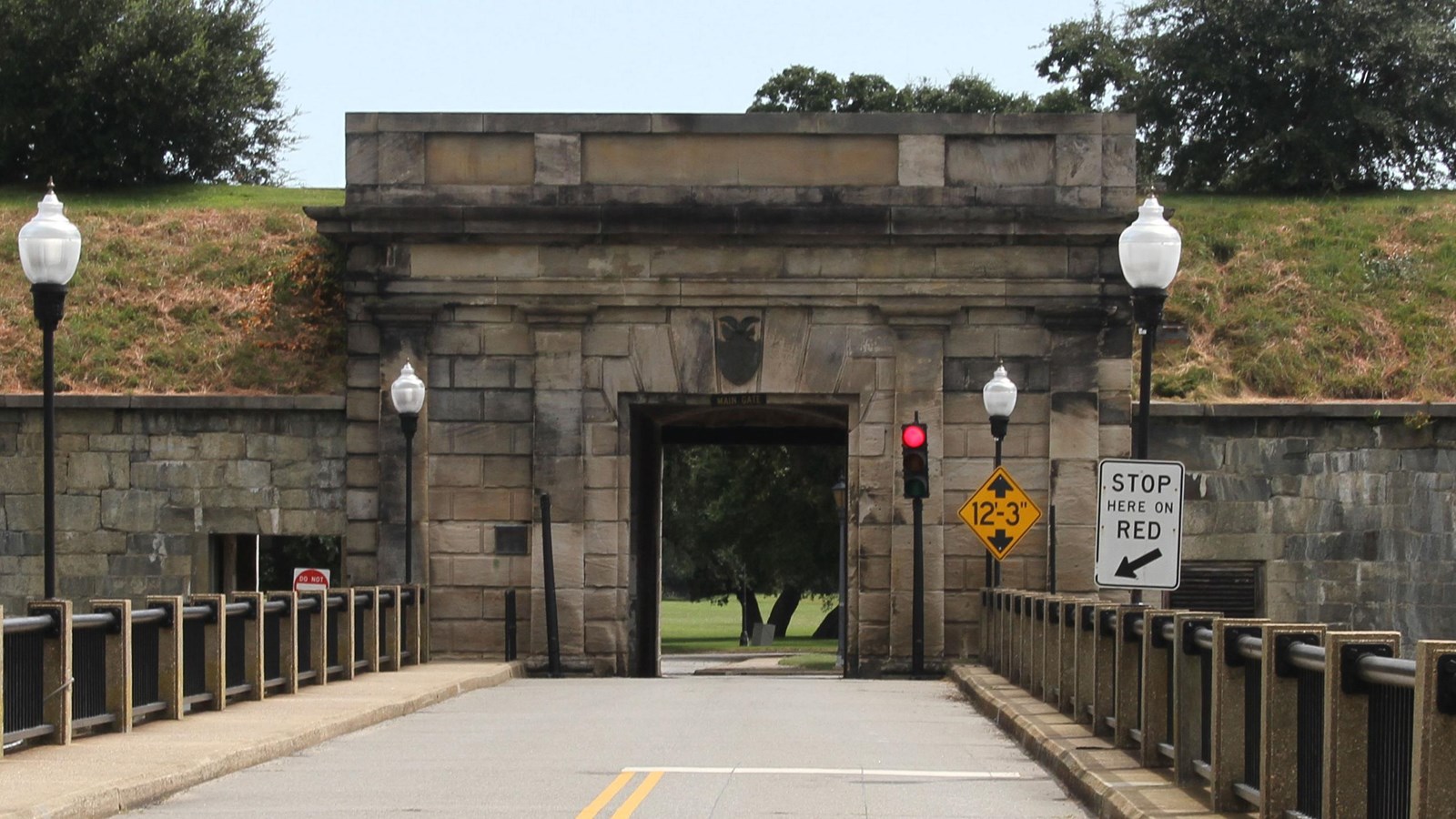 The stone gate of a 1800s fort is approached by a moat bridge with a modern road. 