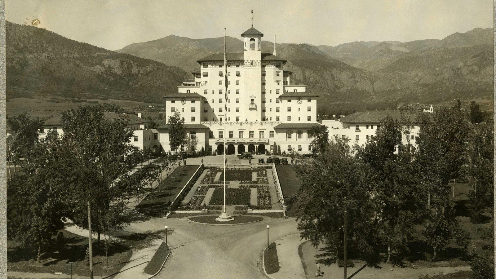 Black and white of large hotel in mountains with grass lined paths leading to it lined by trees. 
