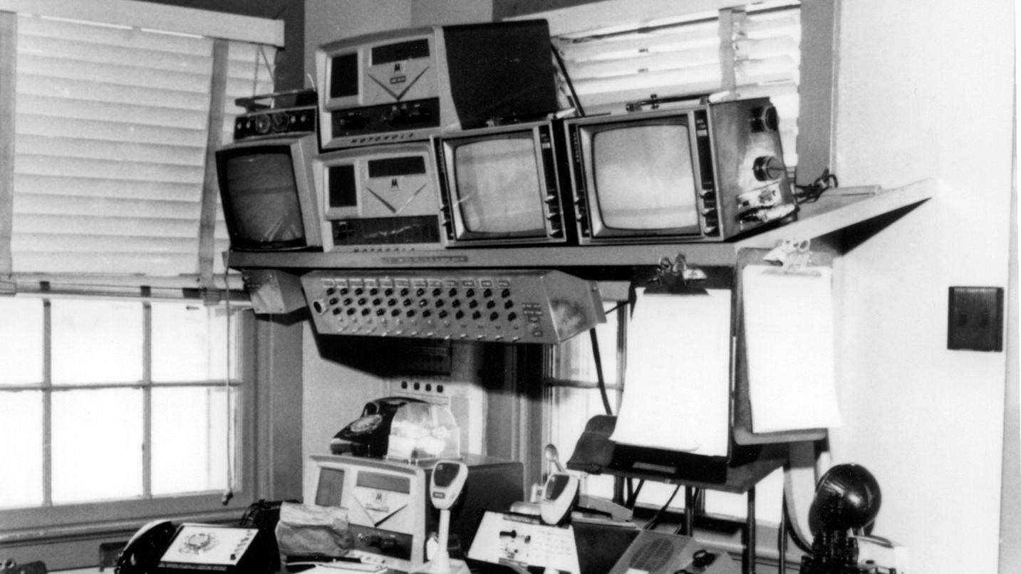 A black and white image of a metal desk with phones, cameras, television screens, and other tech.