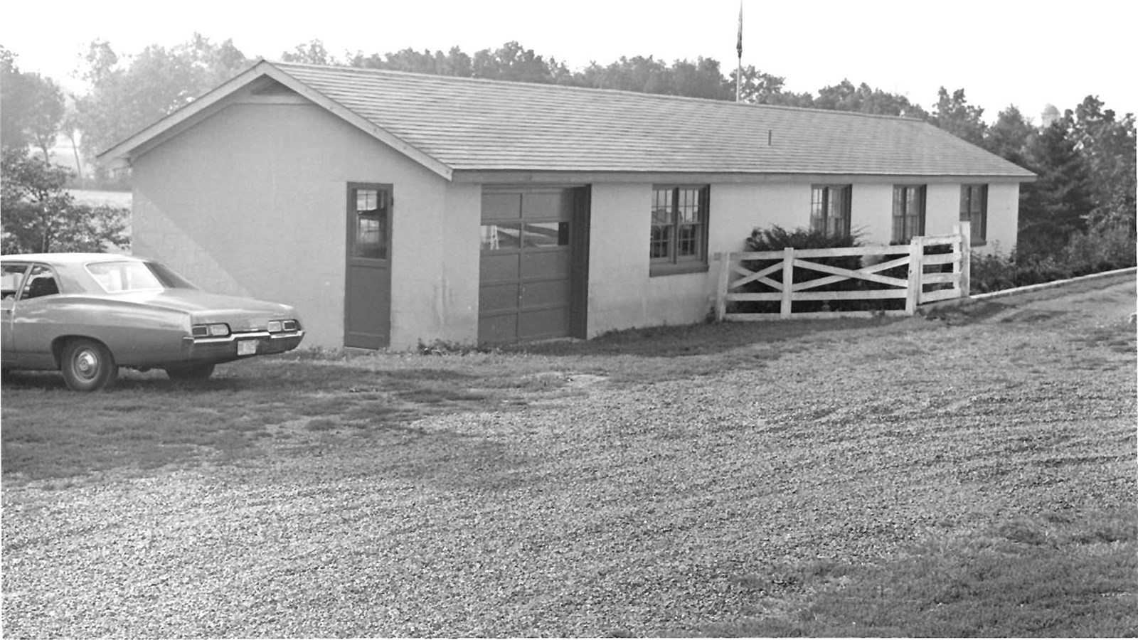 A black and white image showing a long rectangular building, with a 1960’s automobile.