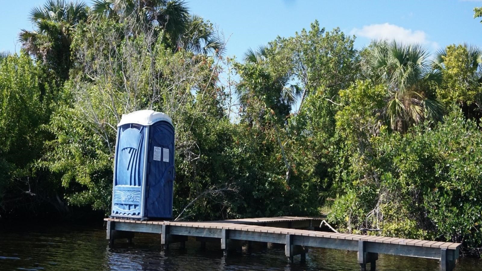 A wooden dock with a blue portable toilet. The dock path bends and disappears into vegetation