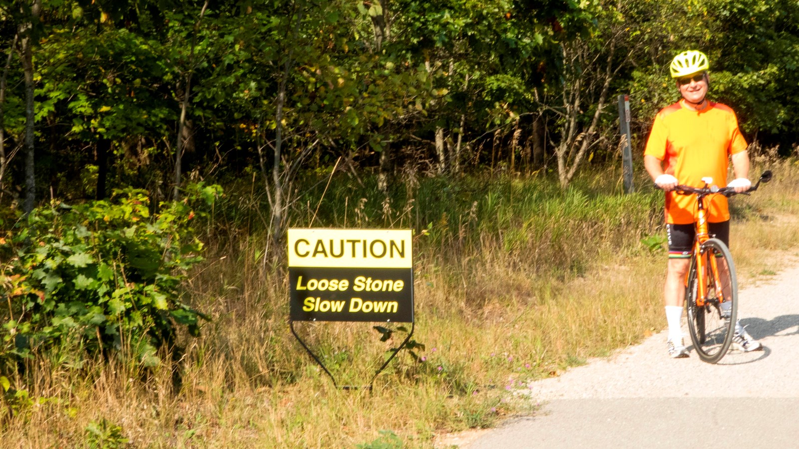 Man with orange bike stands on path near caution sign where paved section meets gravel