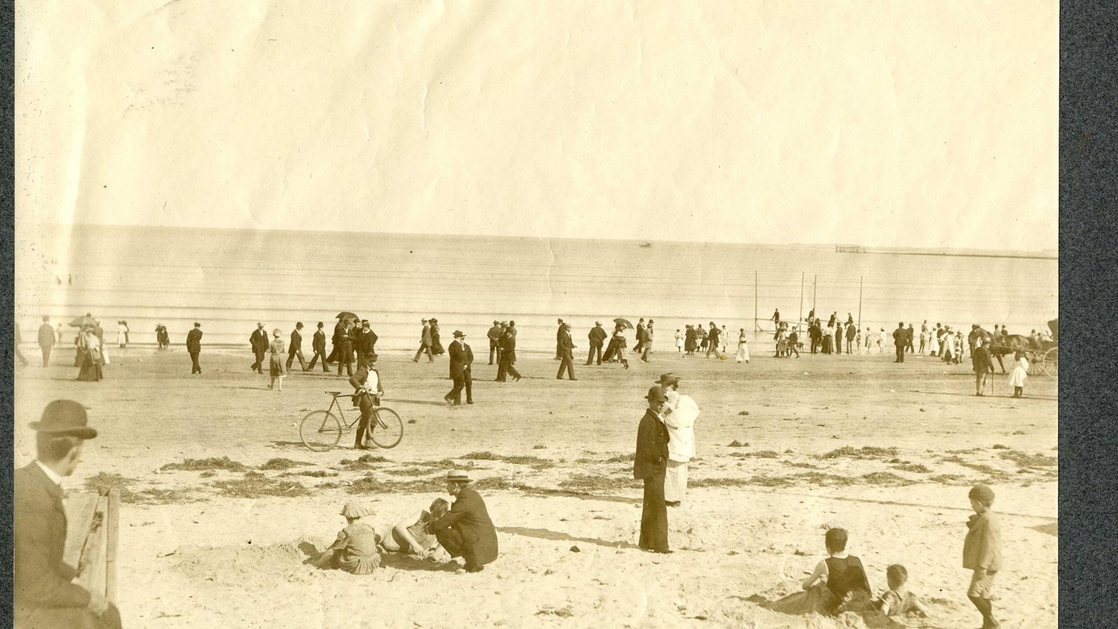 Black and white of sandy area in front of water filled with people, lots wearing suits