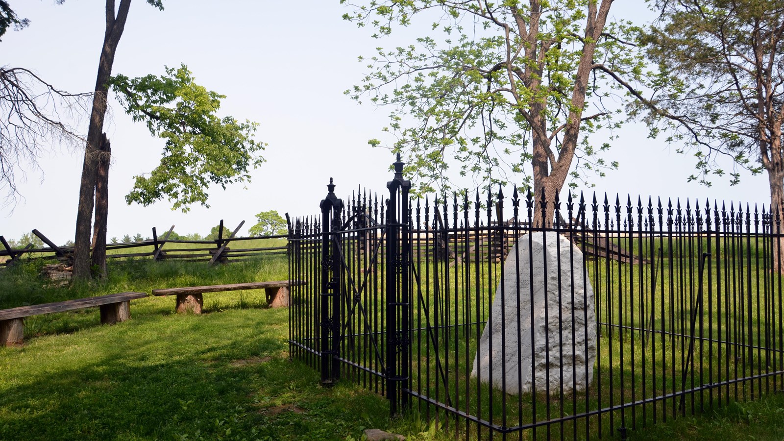 A gated iron fence encloses a marble war memorial in the woods at the edge of a field.
