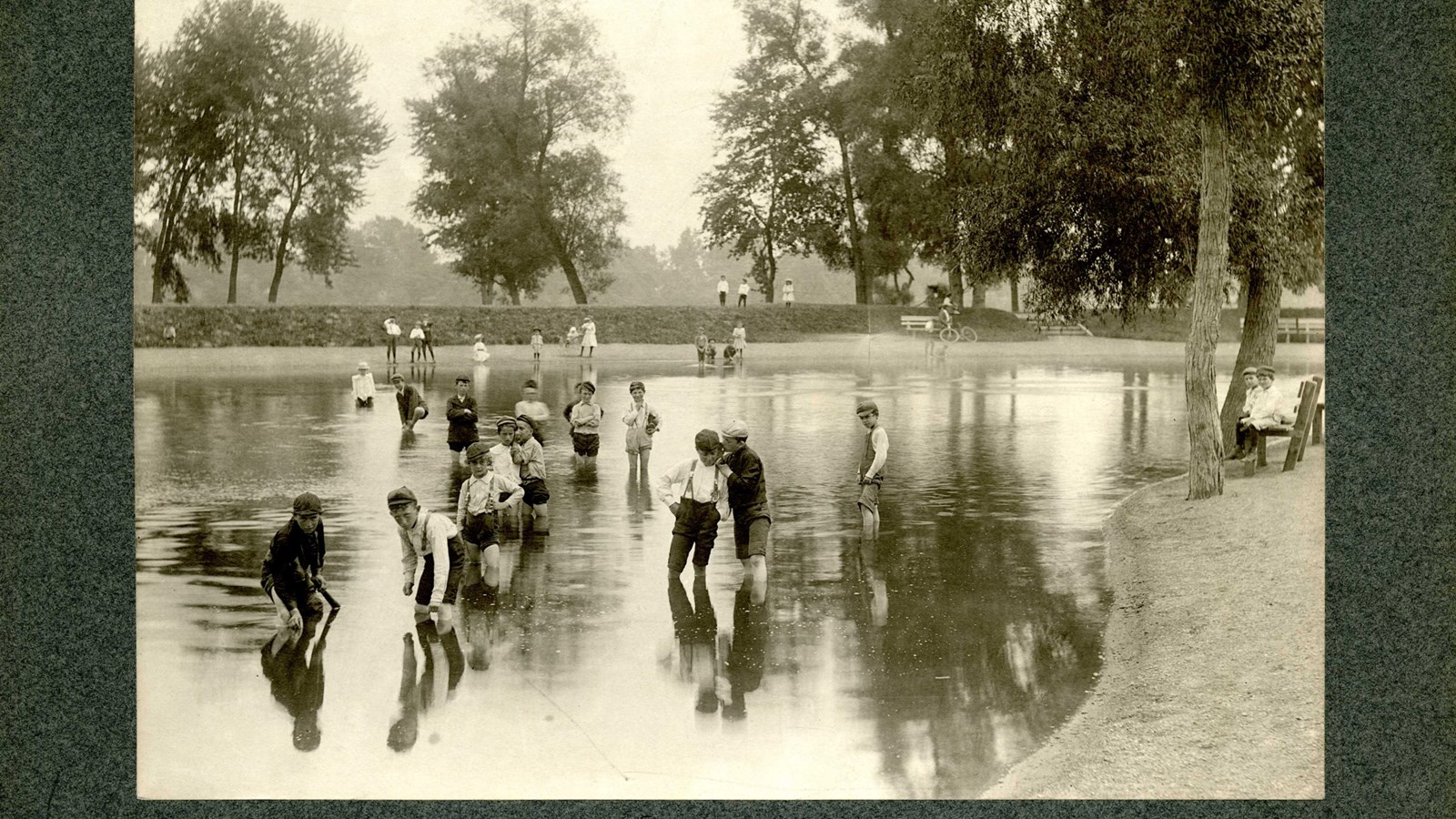 Black and white of sunken body of water with kids playing in it, benches and trees on edge 