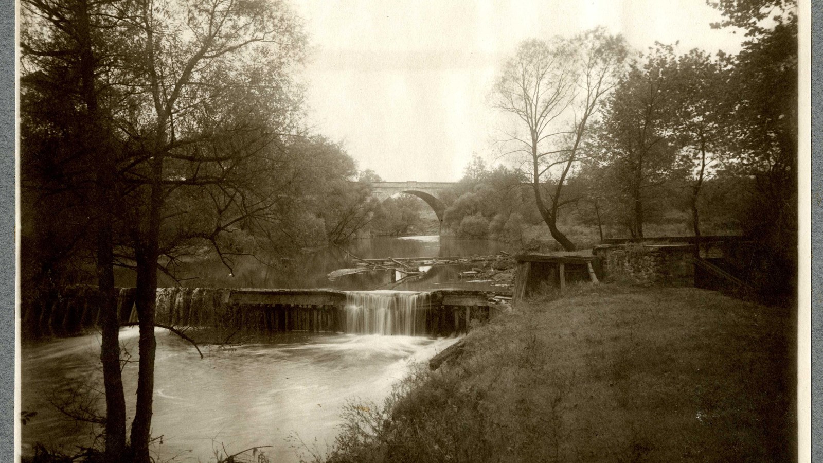Black and white of water running down mini waterfall with water on side, arch bridge in distance