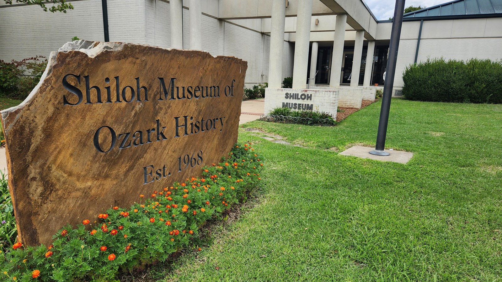 A large stone sign sits in a grassy lawn in front of a one story white building.