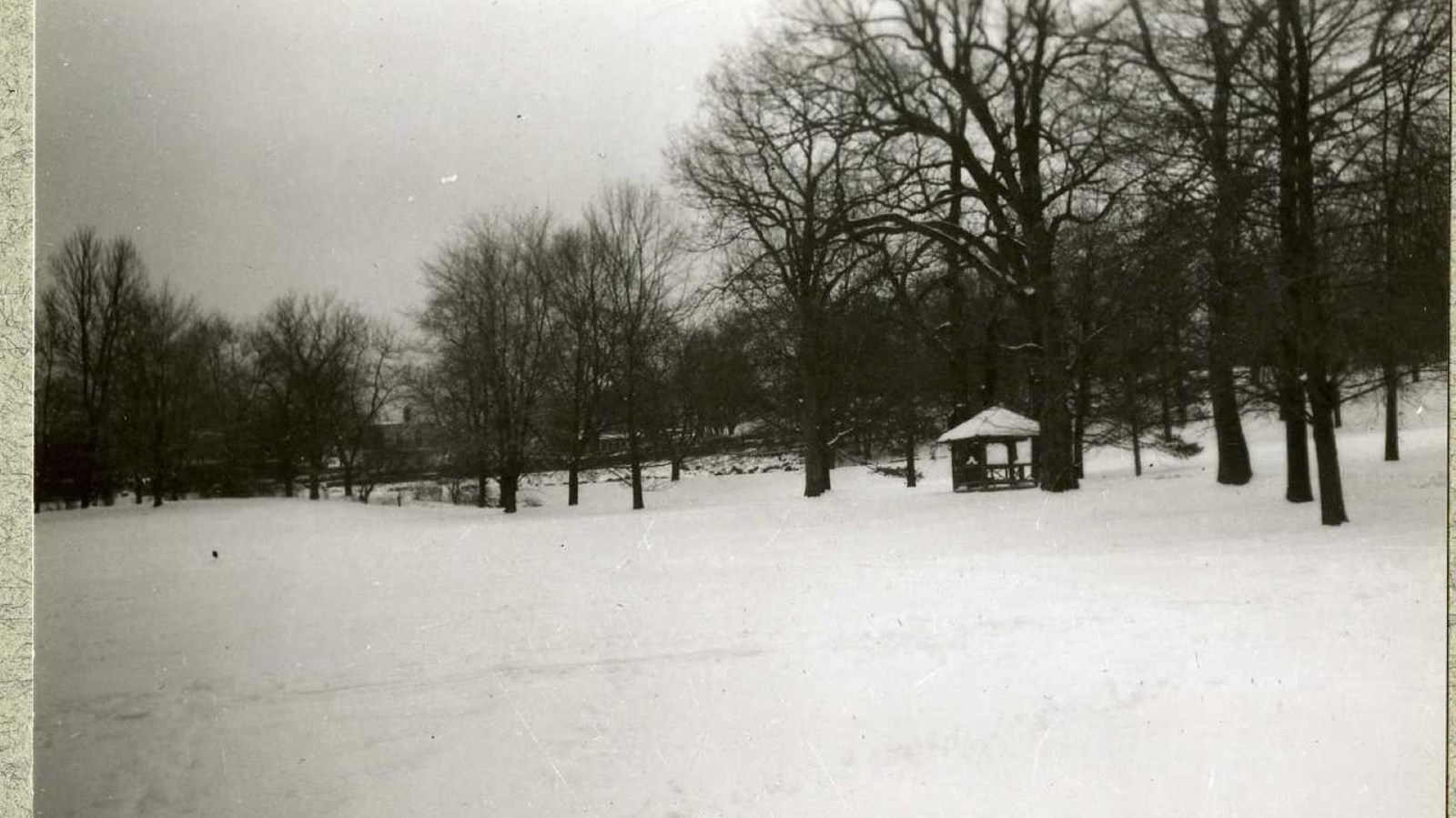 Black and white of flat area with snow on it and shelter in distance with trees around it 