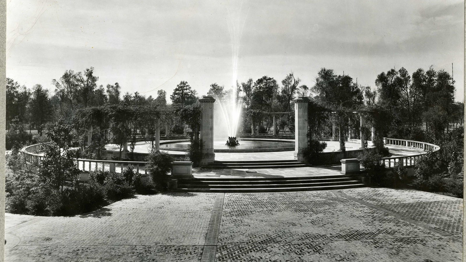 Black and white of large fountain surrounded by trees. 