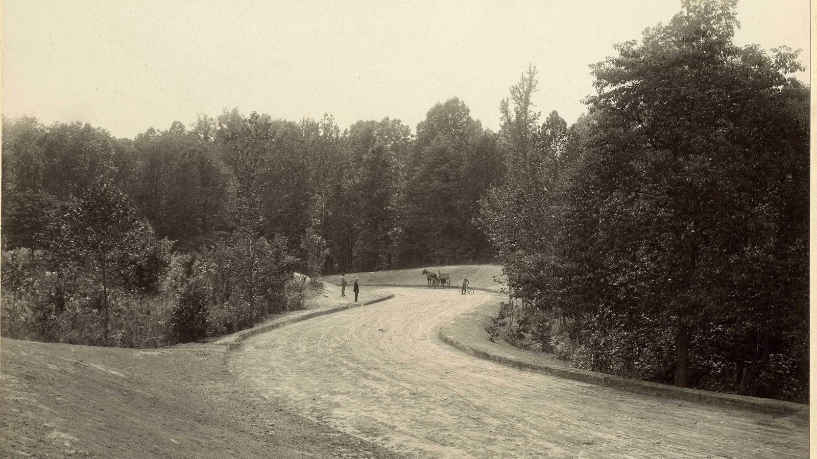 Black and white of curvy dirt path lined with trees on both sides with people and horse and carriage