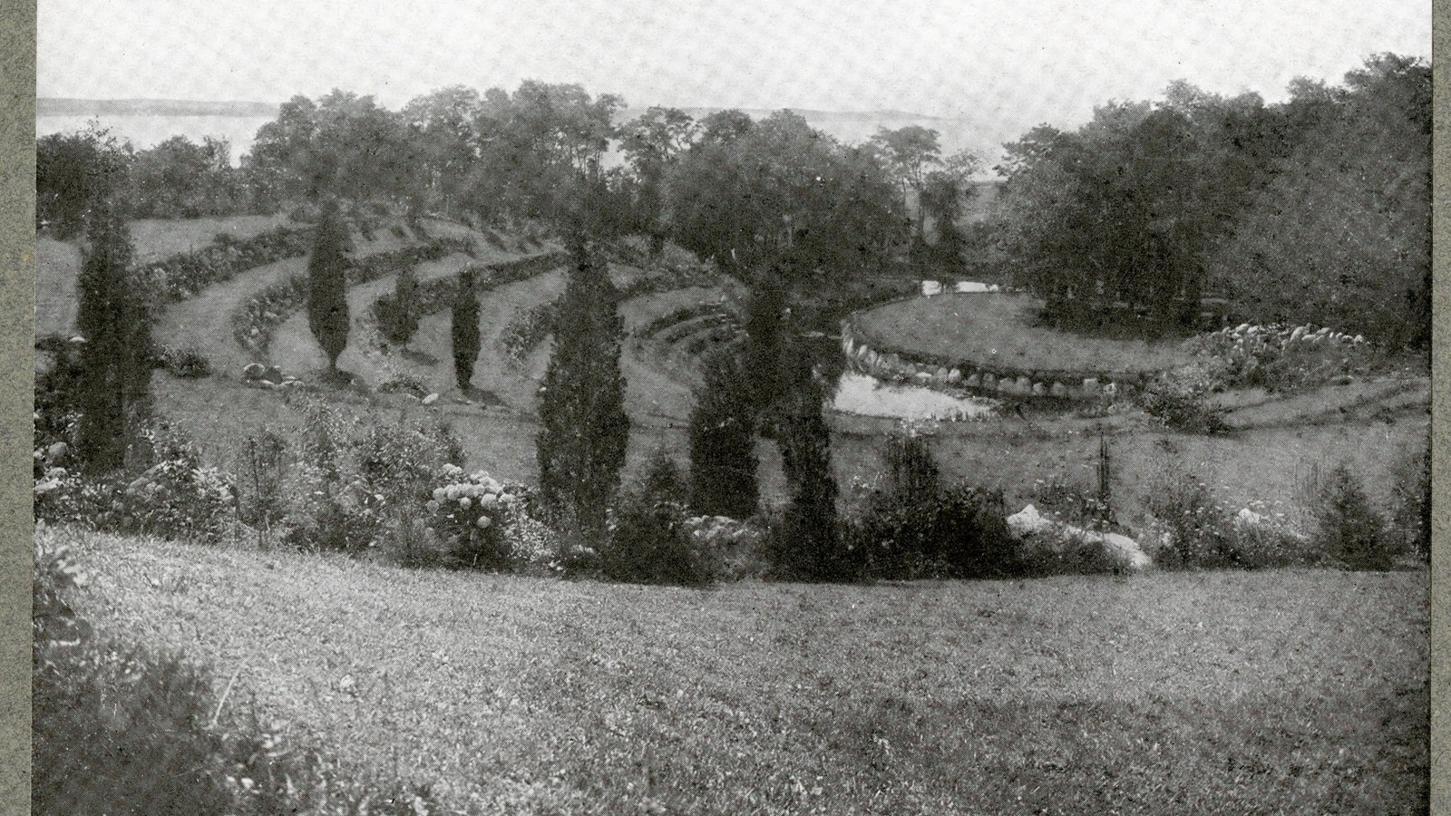 Black and white of hill with stone terraces facing a stage made of grass and rocks. Trees spread out