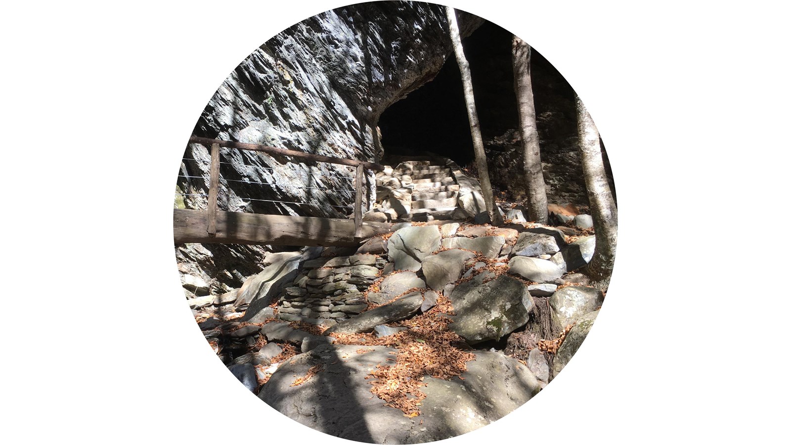 Wooden foot bridge leads to stone stairs that pass under a rocky arch. Rocks and trees nearby.