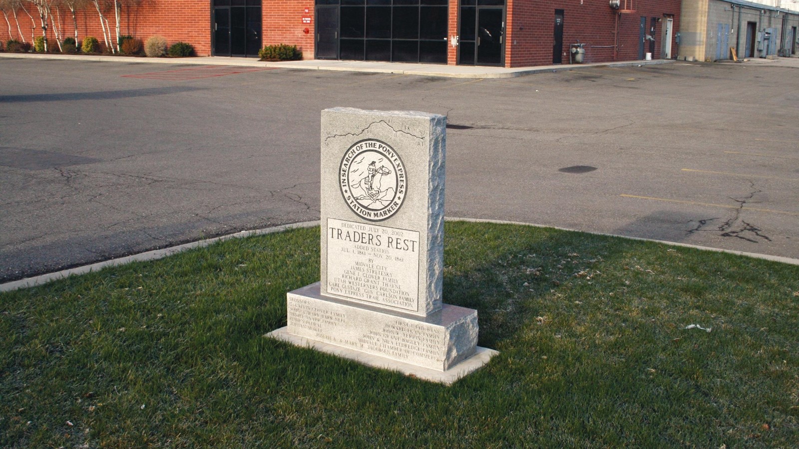 A stone monument with an inscription stands in a grassy corner of a parking lot.