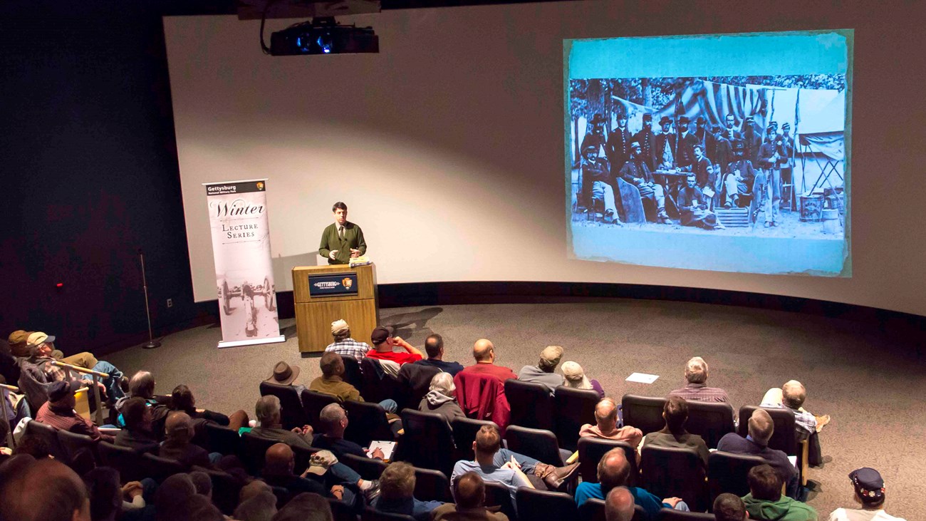 A full theater as seen from the back of the audience. A large white screen, a speaker and podium.