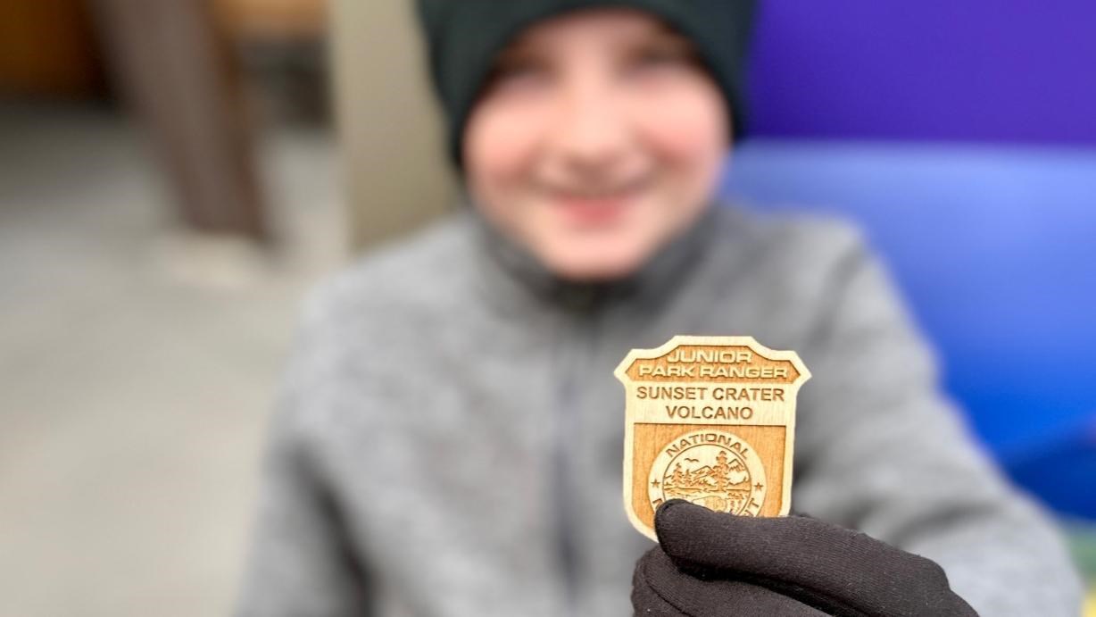 A child wearing cold weather clothing holds up their Jr. Ranger badge. 