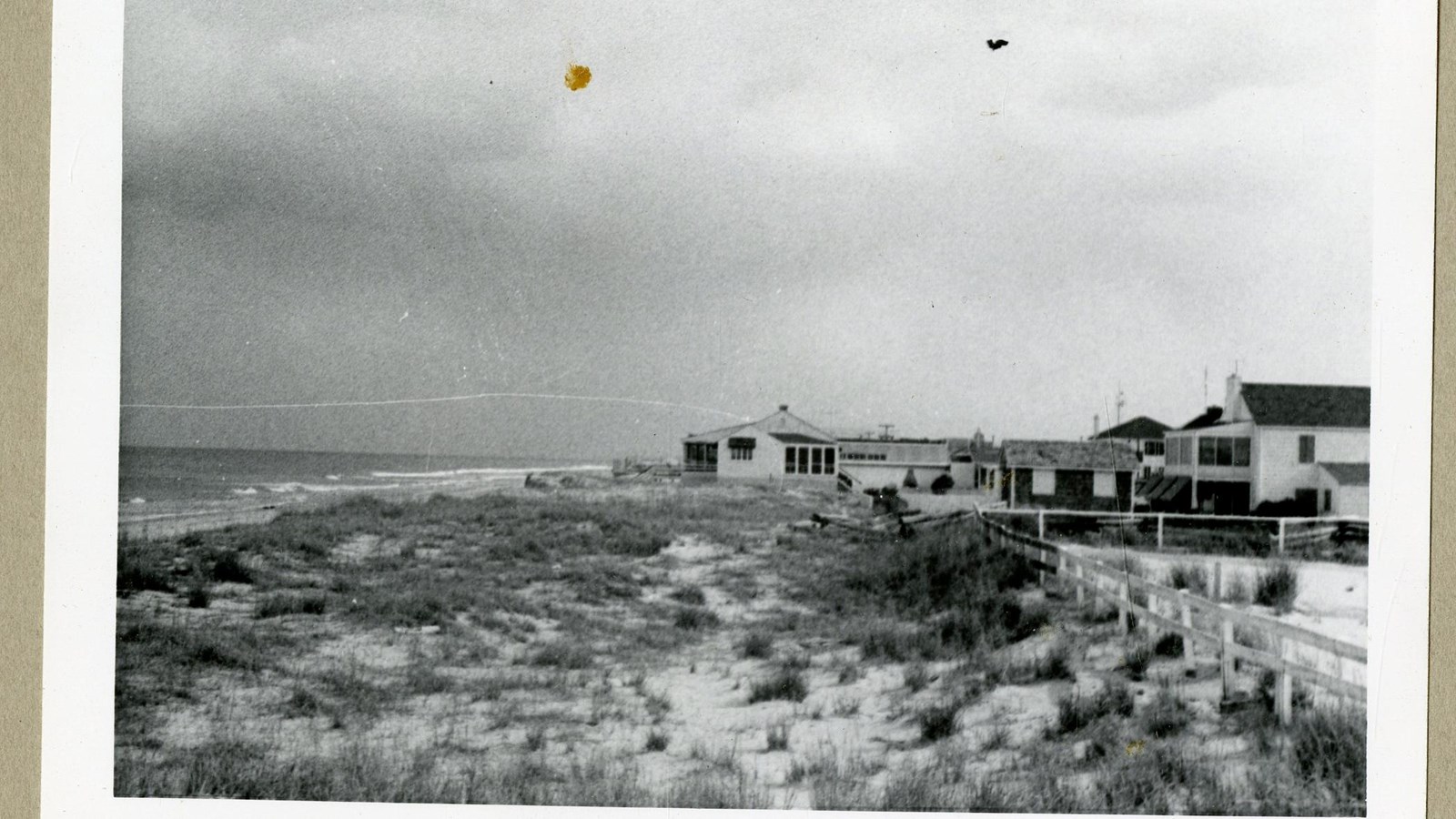 Black and white of sandy area by beach with some grass sticking up, houses behind the sand