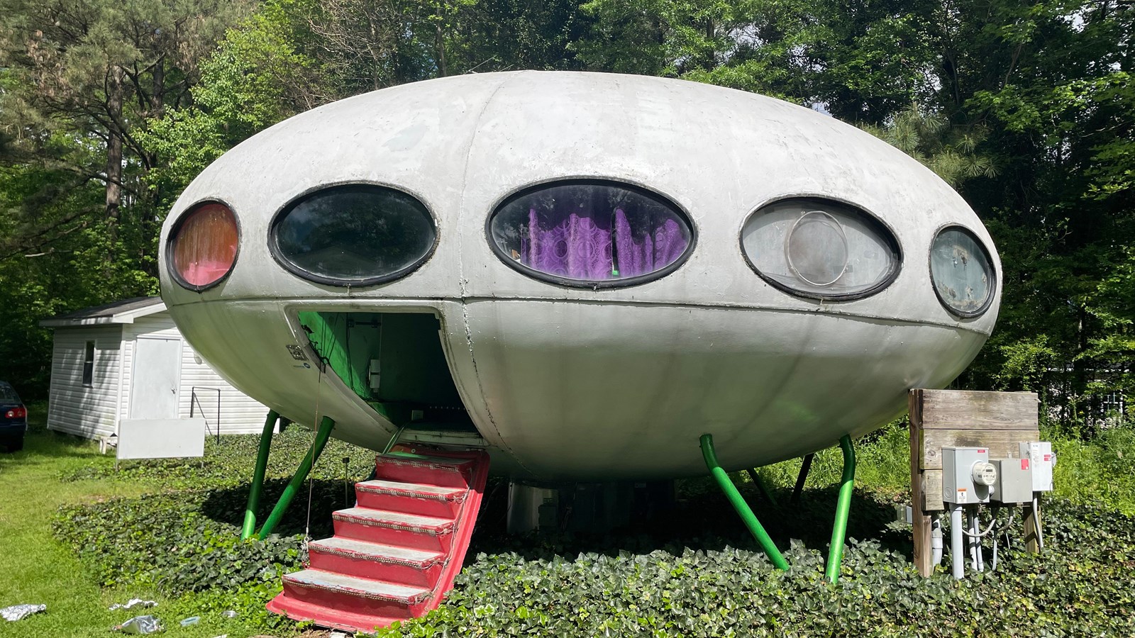 Futuro House, that looks like a landed flying saucer,  set in a green area with trees and a shed 