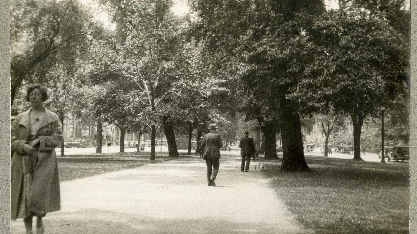 Black and white of flat path with people walking along the path, grass and trees on both sides