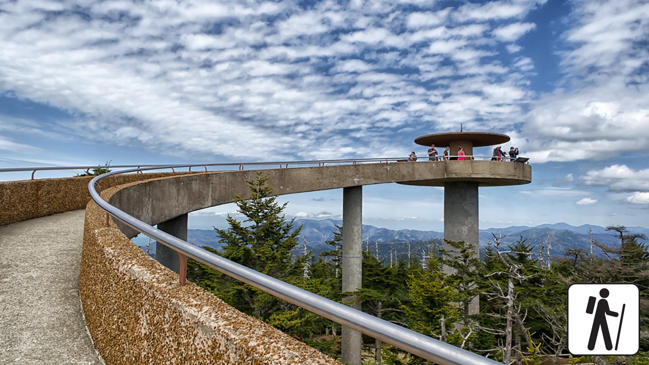 An elevated, paved walkway to a round observation tower overlooking mountains. Hiker icon in corner.