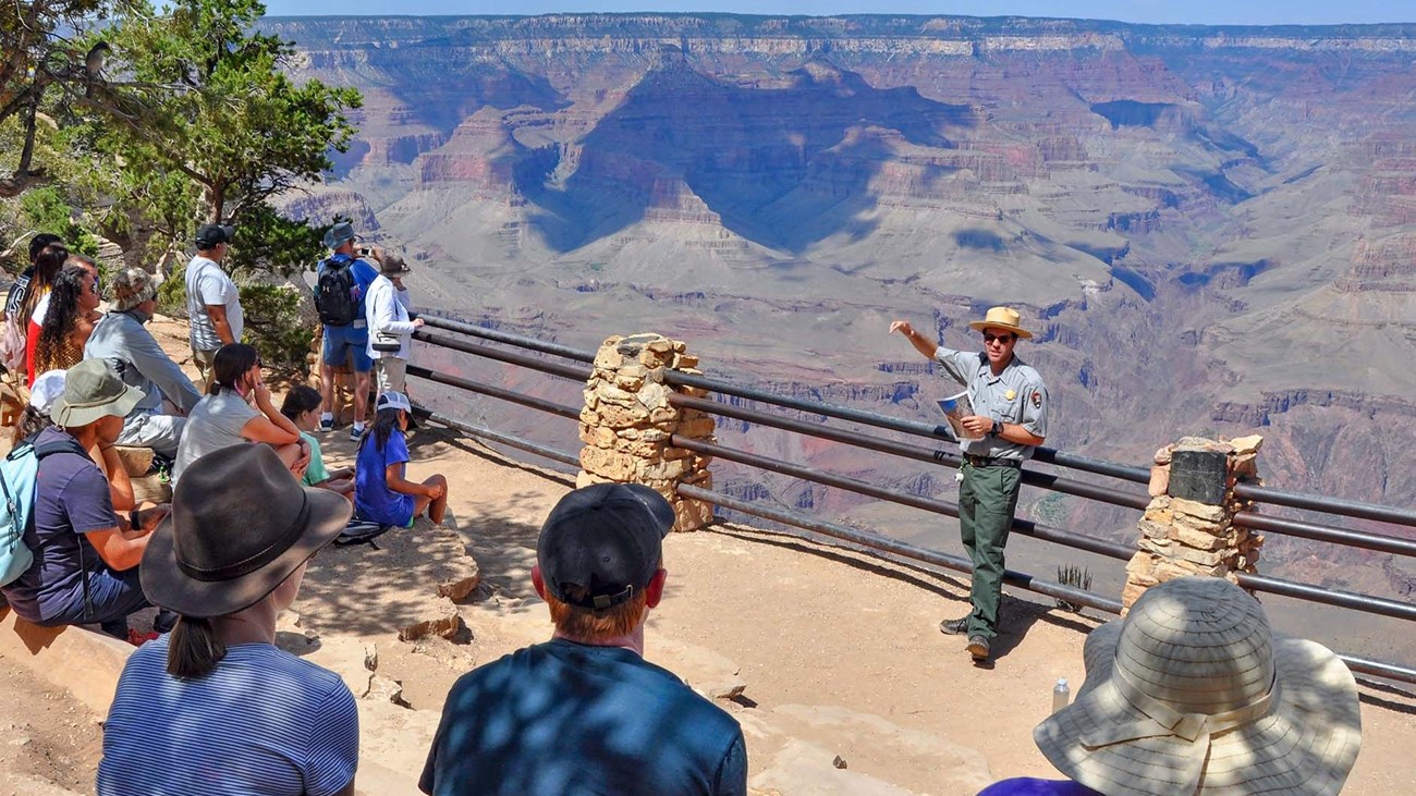 At a scenic overlook with railings, a park ranger is presenting a talk to a group of visitors.