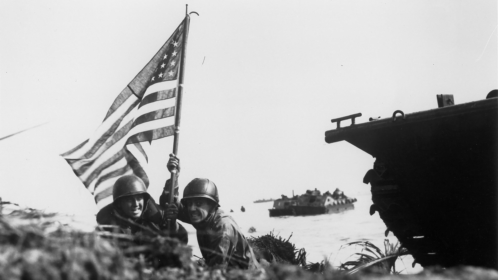 Two soldiers in combat gear crouch next to a tank, holding up a US flag on a boat hook.