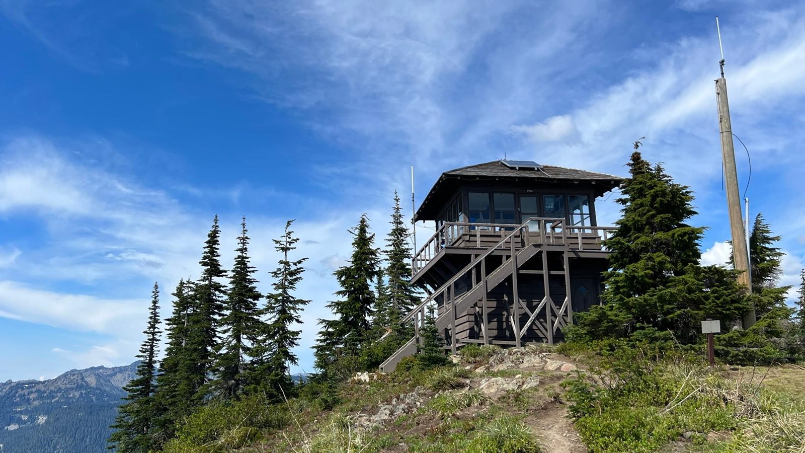 Historic wooden fire lookout painted dark brown sits upon a grassy hillside surrounded by trees