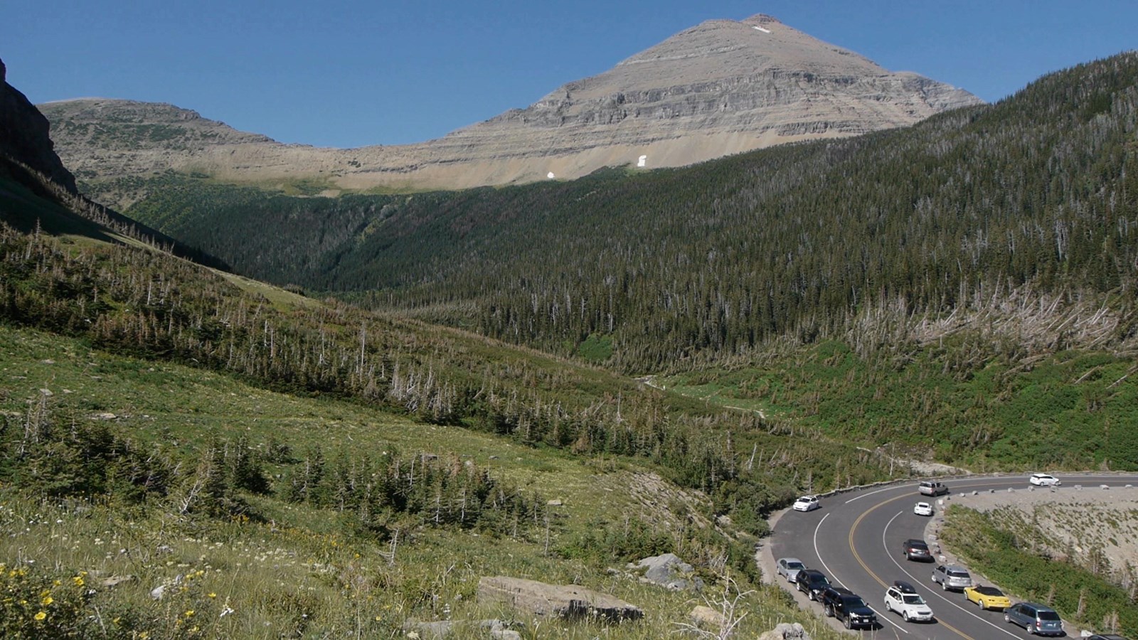 Cars parked along a bend in the road, with a mountain in the distance.