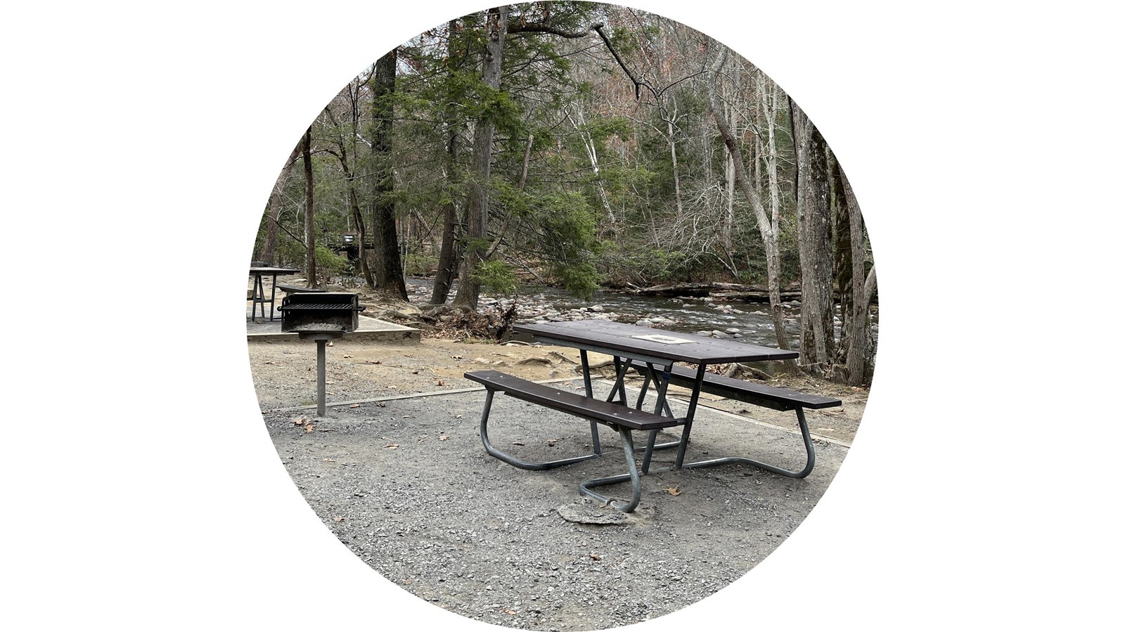 A brown picnic table with metal legs beside a metal grill on a gravel pad along a creek in winter.
