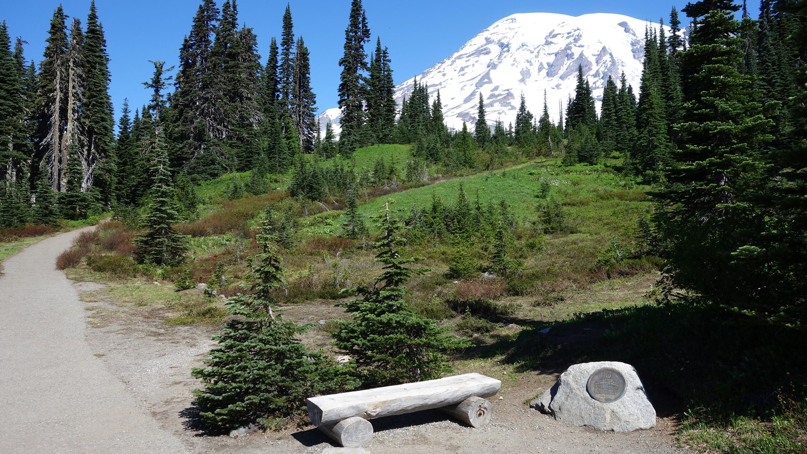 A bronze memorial plaque set into a small boulder next to a trail in front of Mount Rainier.