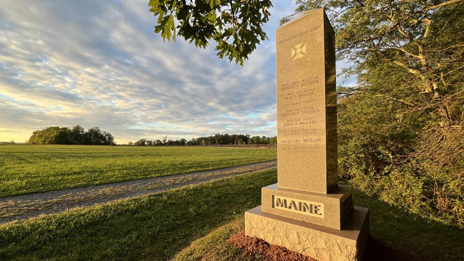 A tall granite obelisk monument on a square base, adjacent to a grassy field. 