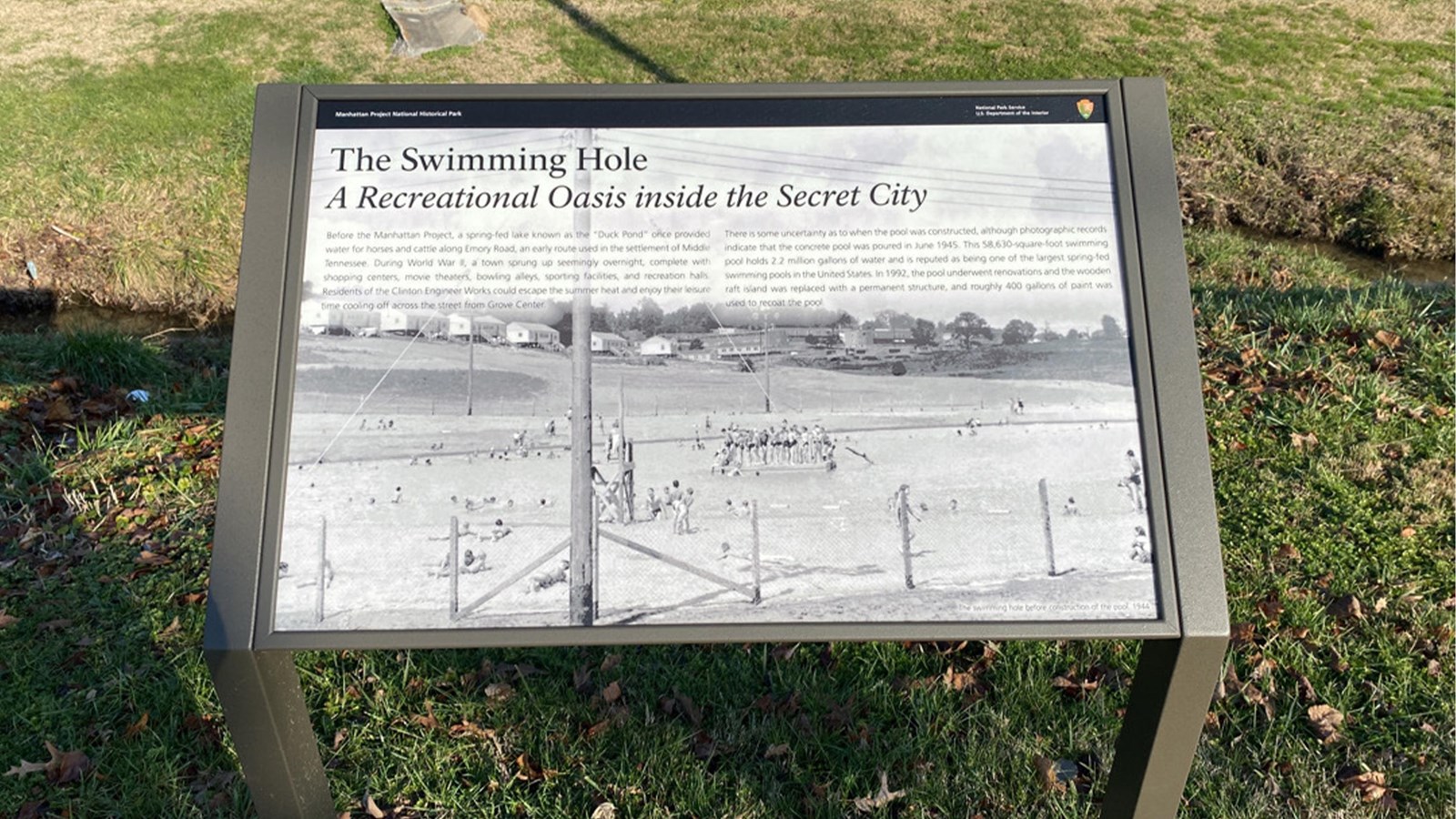 A wayside exhibit on grass in front of a chain link fence.