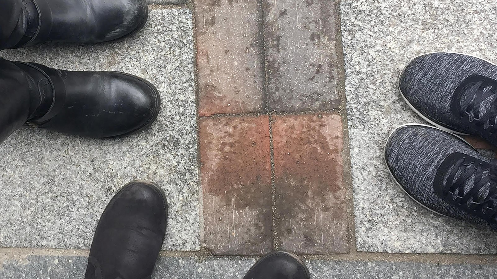 three pairs of feet standing around the start of two lines of red brick on ground.