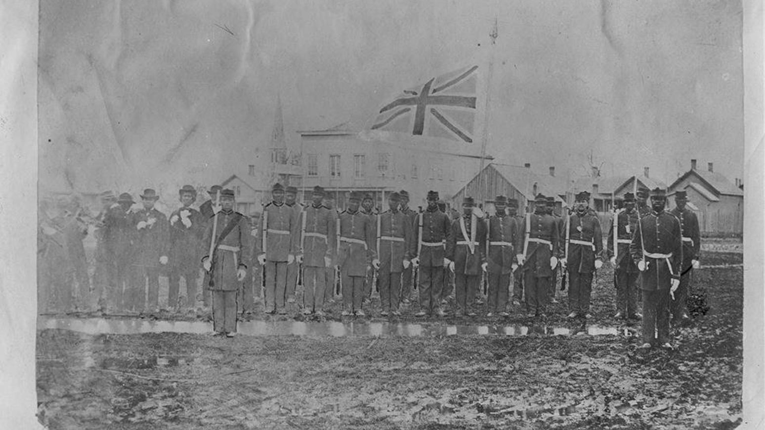 Black and white photograph of men in military uniform on a green with a british flag behind them