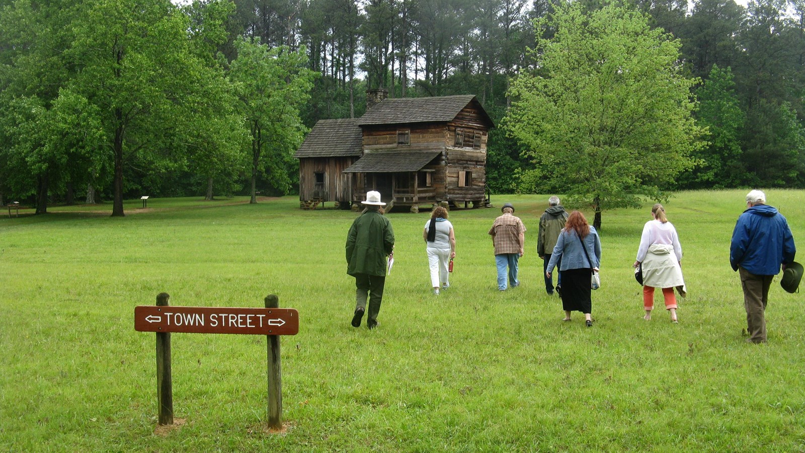 People walk across a grassy field towards a wooden historic building.
