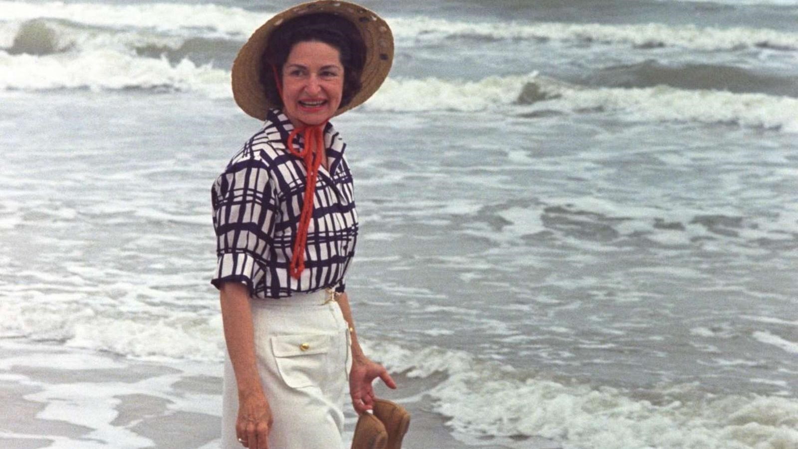 Holding her shoes, Lady Bird Johnson smiles as she walks along the beach in the surf. 